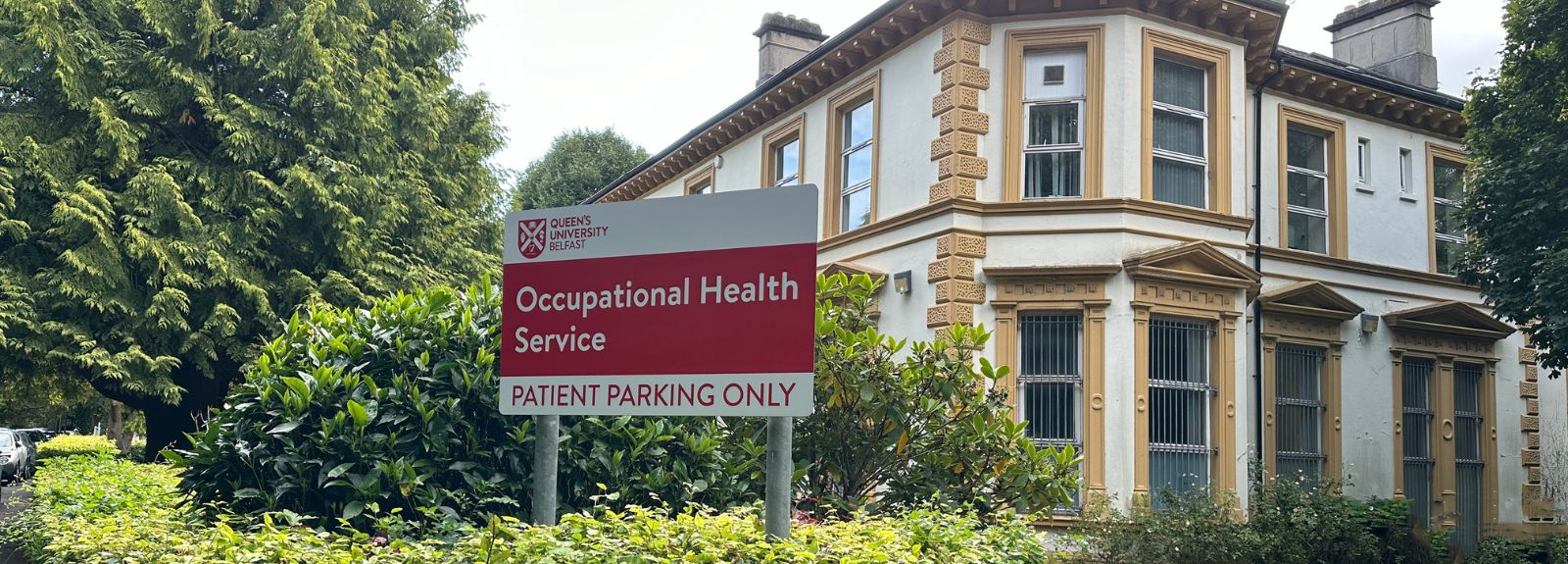 Entrance to 5 Lennoxvale with a sign displaying the QUB logo on white background, with Occupational Health services below written in white on a red background and Patient Parking in red on a white background at the bottom.